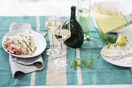 Ein frühligshaft gedeckter Tisch mit gefüllten Weißweingläsern, Bocksbeutelflasche sowie weißen und grünen Spargelstangen.